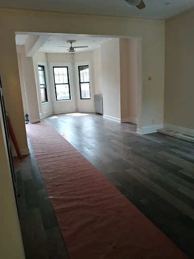 Empty room with cream walls, ceiling fan, three tall windows with black frames, and partially installed hardwood flooring in red and dark tones