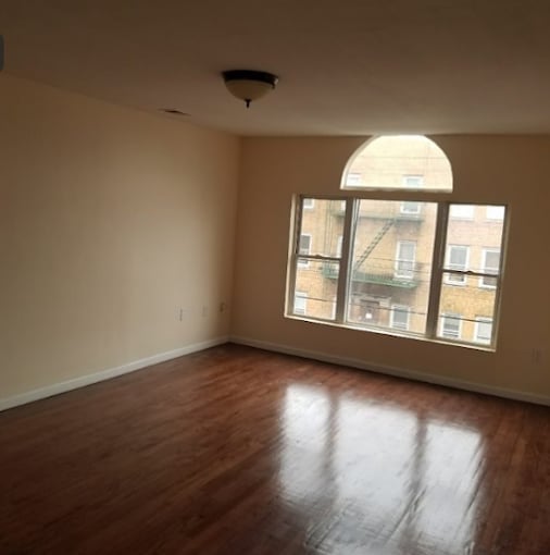 Empty apartment living room with hardwood floors, beige walls, large arched window, and ceiling light fixture