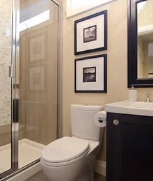 Modern bathroom with beige walls, glass shower enclosure, white toilet, dark vanity, framed artwork, and chrome fixtures