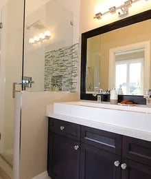 Modern bathroom vanity with dark cabinetry, white countertop, mirror, and shower area with tiled accent wall